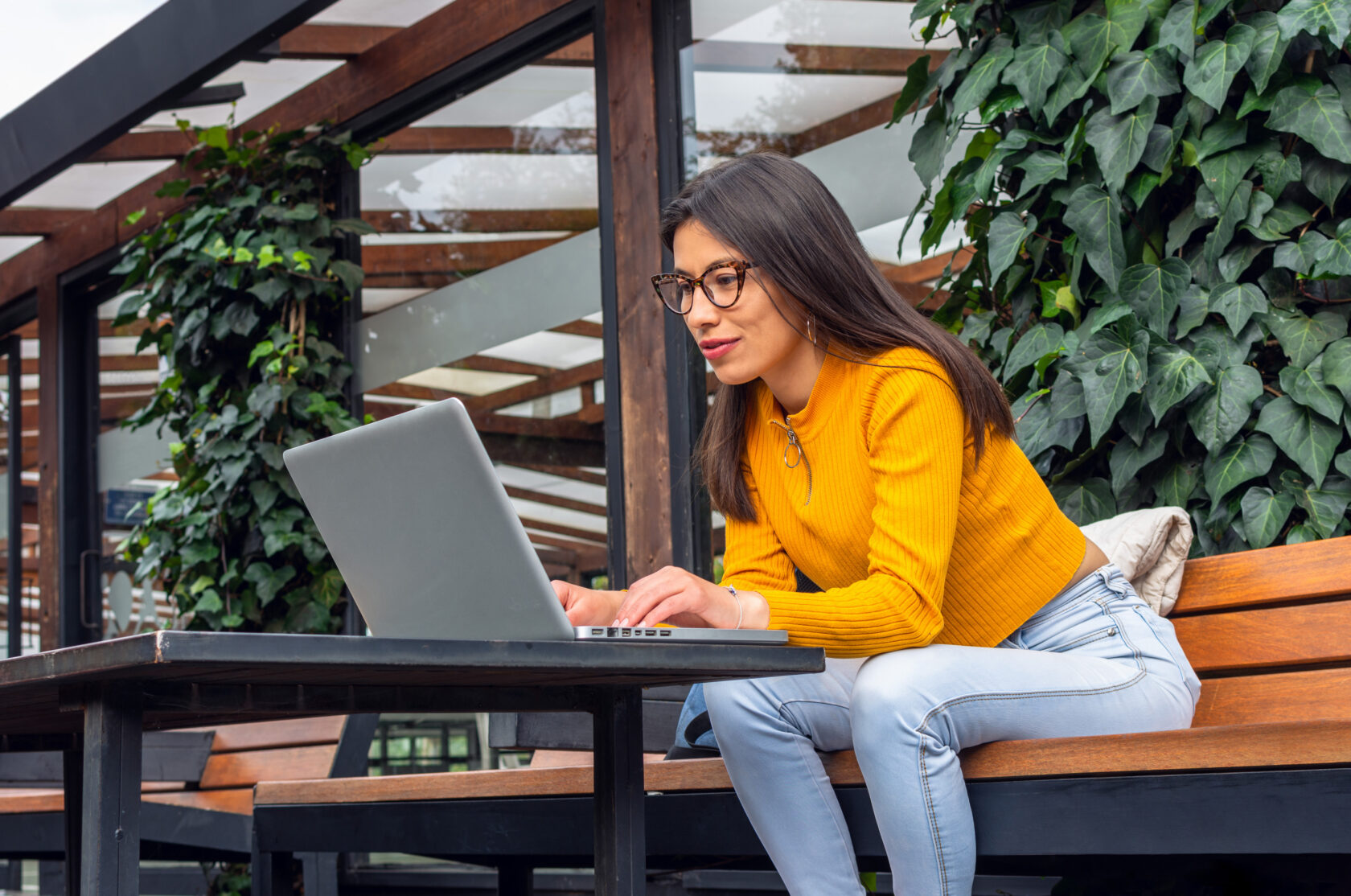 Young, Hispanic woman with indigenous features wearing a yellow sweater sitting on a bench outside while using her laptop